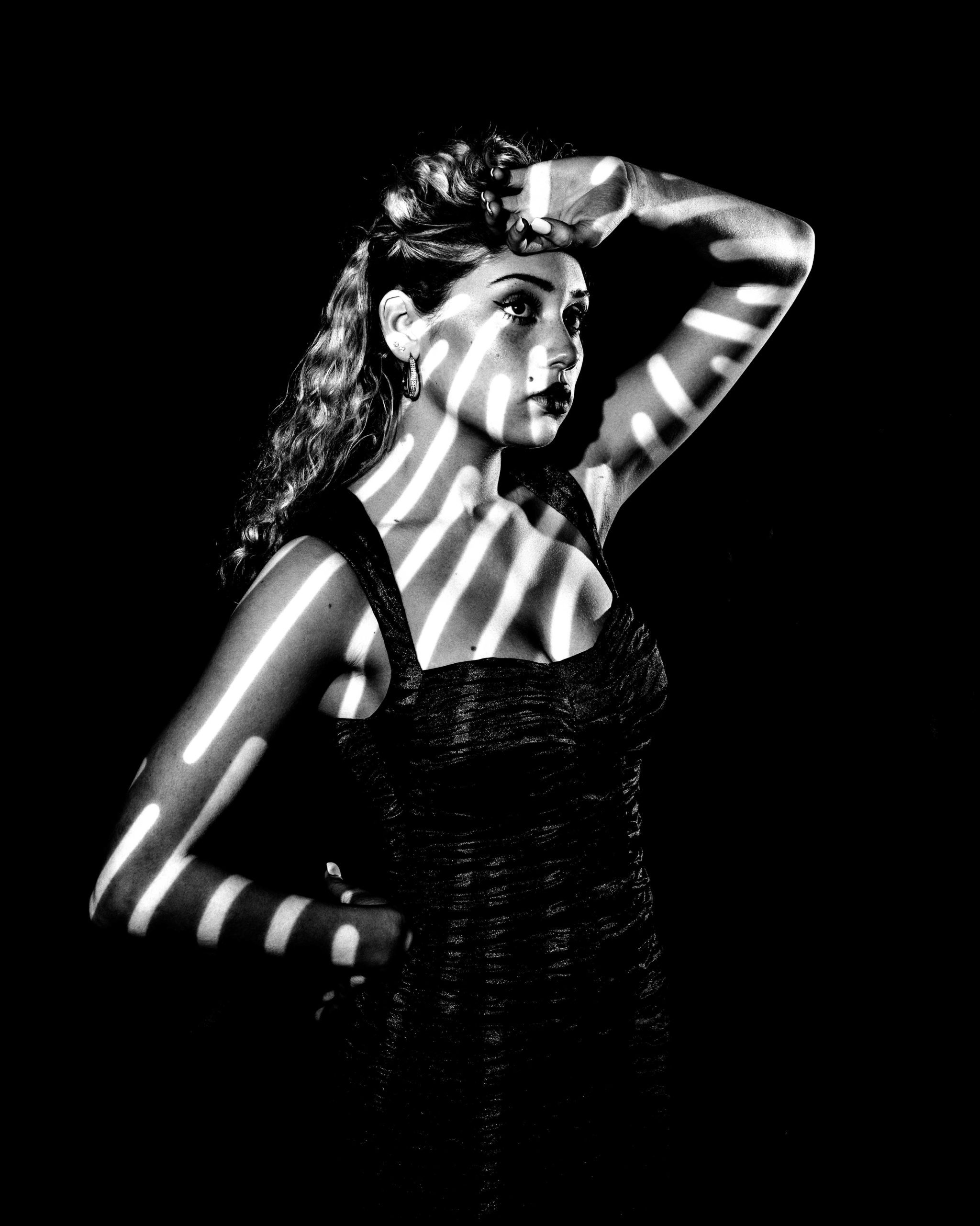 a woman in a black dress is posing with her hands on her head with strips of light illuminating her